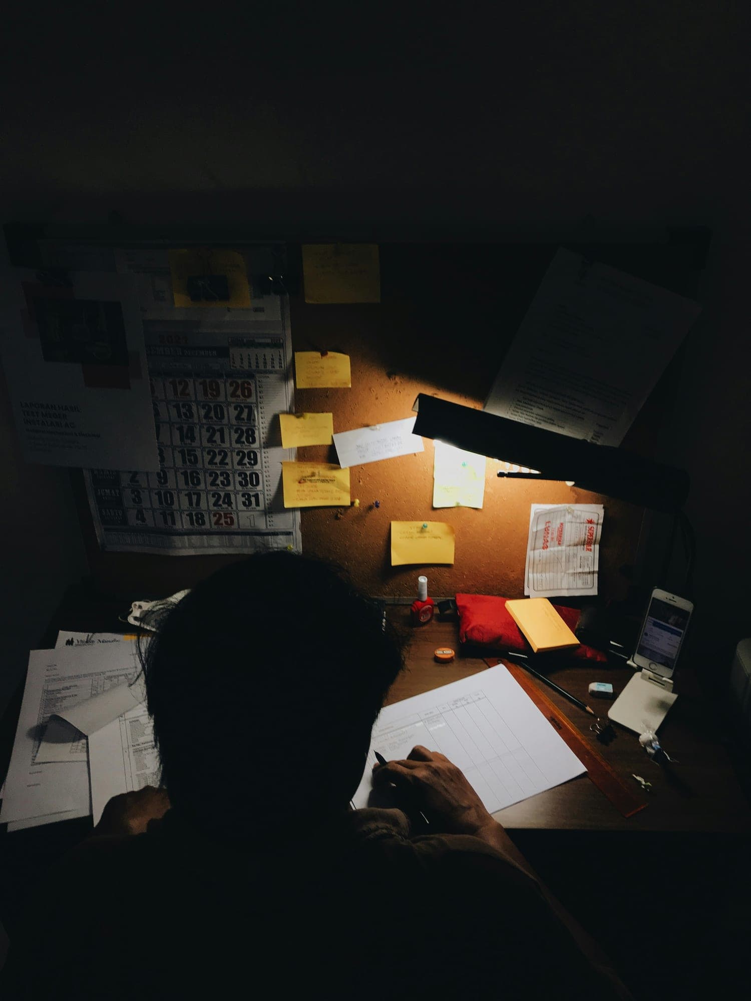 A dim desk lit by a lamp, with notes and a calendar pinned to a corkboard